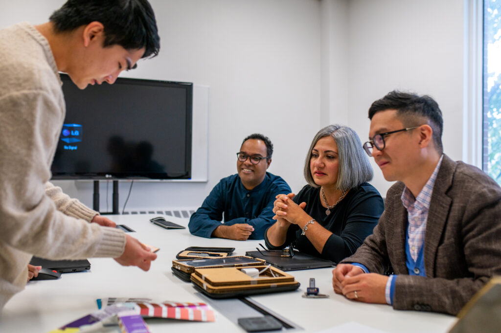 A group of four people in a meeting room engaged in a discussion. Three people sit at a white table — two men and one woman — attentively watching a fourth person standing and demonstrating or presenting something with small objects. The table holds various items, including swatches, a pencil case, and office supplies. A blank TV screen with a "No Signal" message is mounted on the wall in the background, and a large window allows natural light into the room.