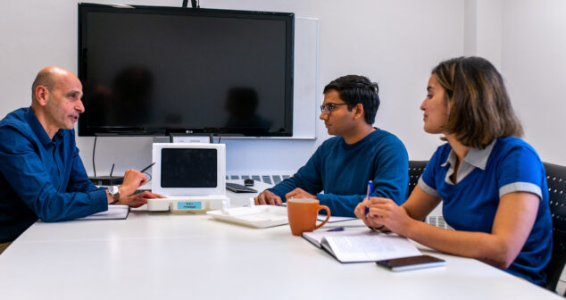 Business Cultures three people talking across a white table with monitor in back.