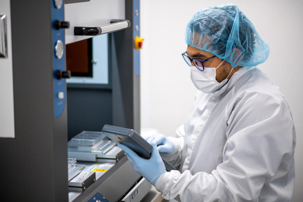 A person working in a cleanroom environment, wearing a white lab coat, blue hairnet, protective face mask, safety glasses, and blue gloves. They are operating or inspecting equipment, holding a tablet or control device, with laboratory machinery and trays visible in the background. The setting appears to be a sterile lab or manufacturing facility.