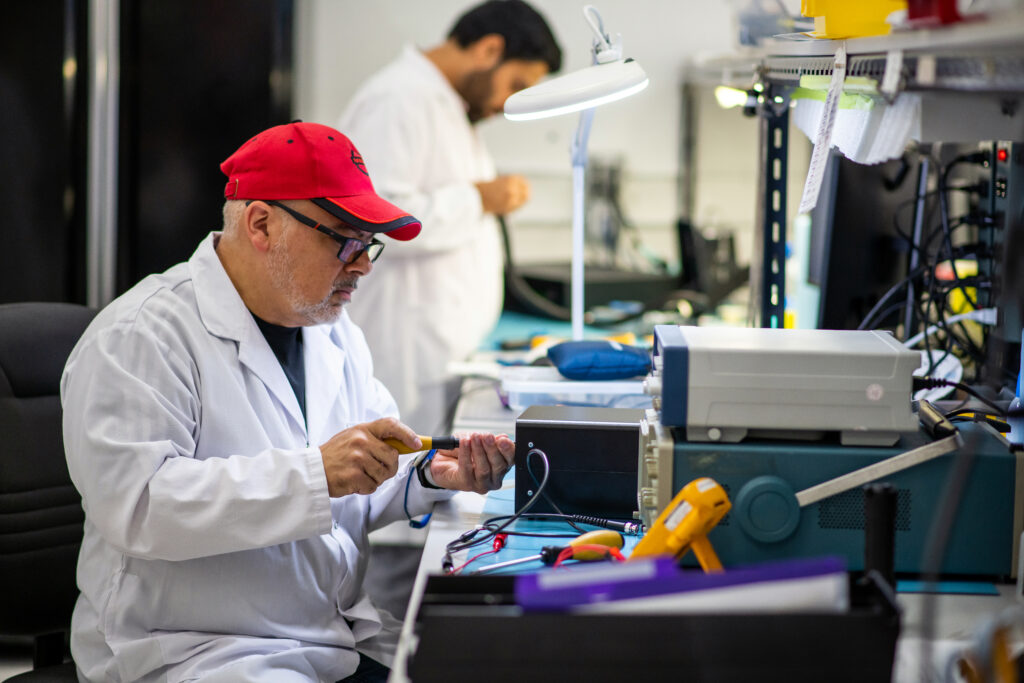 Two technicians working in a laboratory or electronics workshop. In the foreground, a man wearing a white lab coat, red baseball cap, and glasses is seated at a workbench, using a tool to work on an electronic device. The bench is cluttered with various instruments, tools, and equipment. In the background, another technician in a white lab coat is standing and working at a separate station under a bright task light. The environment is organized and technical, indicating detailed assembly or testing work.