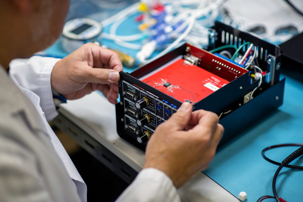 The image shows a close-up of a person working on an electronic device, likely a piece of testing or medical equipment. The device's casing is open, revealing internal components such as circuit boards, wiring, and connectors. The individual, dressed in a white lab coat, is carefully handling a panel, which includes knobs and input/output ports. The background features a workspace with tools and wires, indicating a technical or engineering environment focused on device assembly, testing, or maintenance.