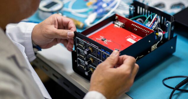 The image shows a close-up of a person working on an electronic device, likely a piece of testing or medical equipment. The device's casing is open, revealing internal components such as circuit boards, wiring, and connectors. The individual, dressed in a white lab coat, is carefully handling a panel, which includes knobs and input/output ports. The background features a workspace with tools and wires, indicating a technical or engineering environment focused on device assembly, testing, or maintenance.