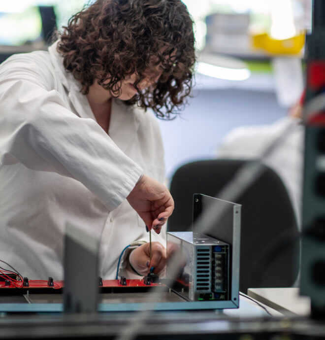 The image shows a person with curly hair wearing a white lab coat working on an open electronic device at a workstation. They are using a tool, such as a screwdriver, to adjust or assemble components on a circuit board. The device appears to be partially assembled, with visible wiring and metal panels. The background includes other workstations and equipment, indicating a laboratory or engineering environment focused on device assembly, testing, or prototyping.