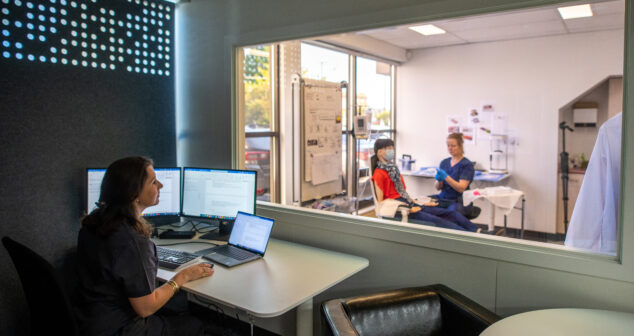 The image shows a professional workspace divided by a glass window. In the foreground, a person is seated at a desk with multiple computer monitors and a laptop, seemingly observing or recording data. Through the glass window, two individuals are visible in a clinical or lab-like setting. One is seated in a chair, wearing a mask and scarf, while the other, dressed in scrubs and gloves, appears to be conducting a procedure or simulation. The environment suggests a usability testing scenario.