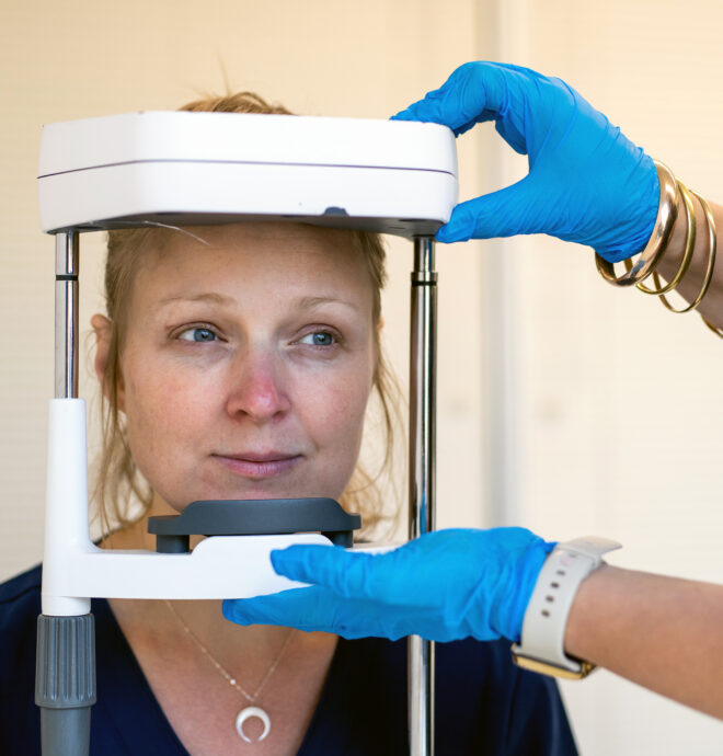 The image shows a close-up of a woman seated, with her head positioned in a medical or diagnostic device. The device appears to be a headrest or stabilizer used for imaging or examination purposes. A person's hands, wearing blue gloves, are seen adjusting the device. The setting suggests a clinical or research environment, emphasizing precision and care in handling the equipment. The woman appears calm and cooperative during the procedure.