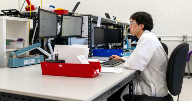 The image shows a person wearing safety goggles and a lab coat working at a desk in a laboratory or engineering workspace. They are seated at a computer station with multiple monitors, typing on a keyboard while focusing on the screen. The desk is equipped with tools, documents, and a red organizer tray. The background features shelves and technical equipment, indicating a controlled, professional environment for research, development, or testing purposes.