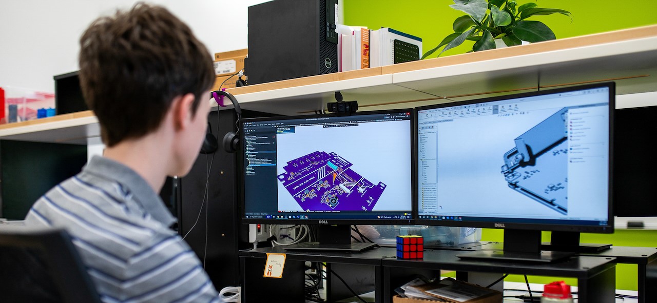A young person wearing a striped shirt is seated at a desk with dual monitors. They are working on computer-aided design (CAD) software, displaying a 3D model of a purple printed circuit board (PCB) on the left screen and a mechanical CAD rendering of a device enclosure on the right screen. The workspace includes a webcam mounted between the monitors, a Rubik’s Cube, a headset, and some office supplies. The background features a green wall and a shelf with books and a potted plant.