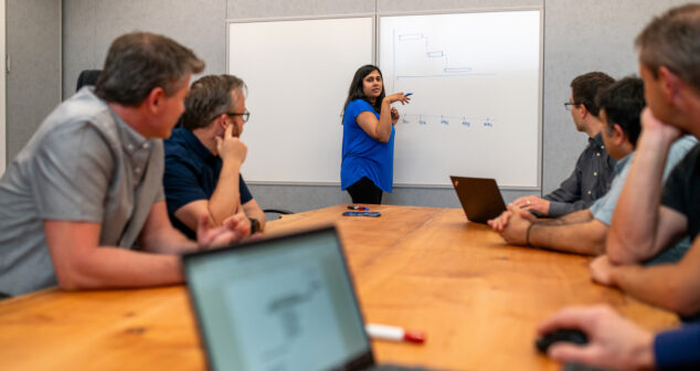 A team of professionals is seated around a conference table while a woman in a blue blouse stands at the whiteboard, explaining a diagram. The table has laptops and papers, and the atmosphere appears collaborative and focused.