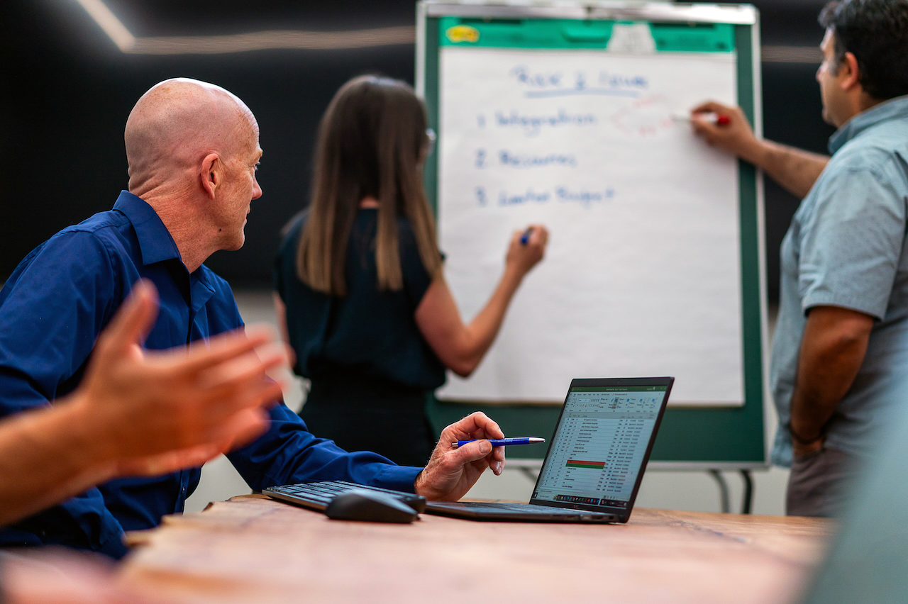 Three people engaged in a collaborative meeting. In the foreground, a man in a blue shirt sits at a table with a laptop showing a spreadsheet, gesturing with a pen in hand. In the background, a woman and another man stand in front of a large flip chart, writing notes. The flip chart has headings and bullet points written in blue marker. The setting appears to be a modern office or meeting space.