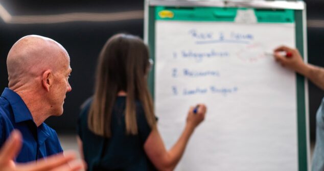 The image depicts a group of people engaged in a collaborative discussion or brainstorming session. A woman is writing on a flip chart with a marker, while a man on her right is pointing at the chart with a marker, and another man on her left is attentively observing. The flip chart includes a list of numbered topics under the heading "Risk & Issues." The setting appears to be a professional or corporate environment.