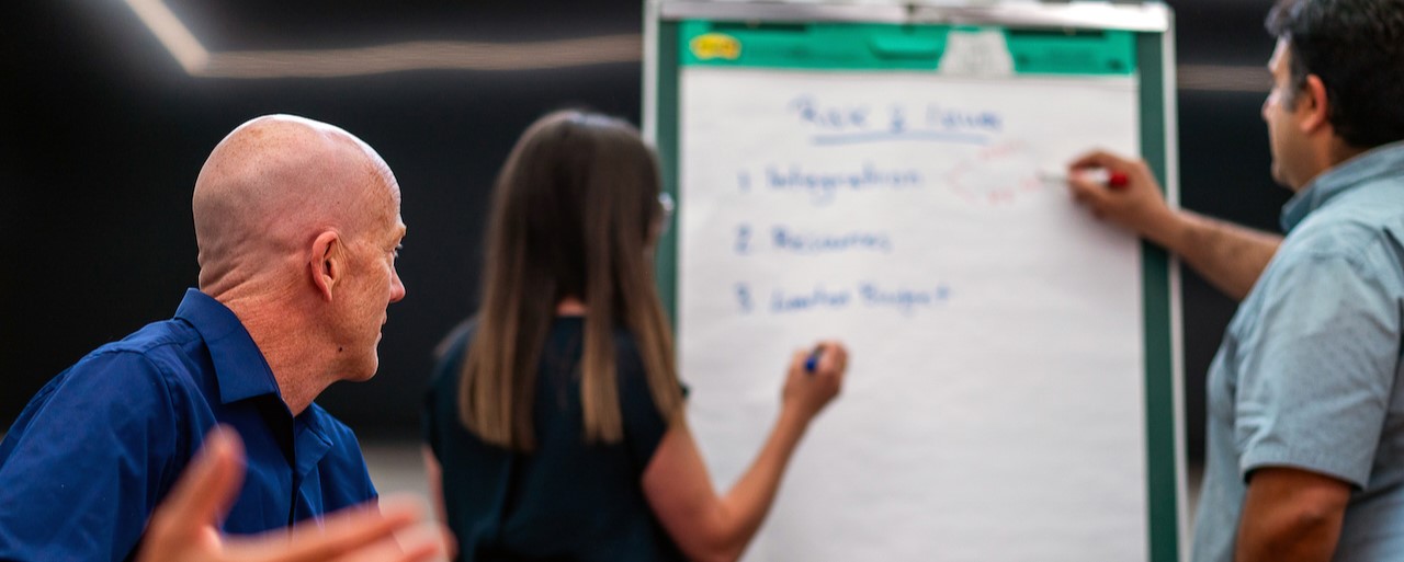 The image depicts a group of people engaged in a collaborative discussion or brainstorming session. A woman is writing on a flip chart with a marker, while a man on her right is pointing at the chart with a marker, and another man on her left is attentively observing. The flip chart includes a list of numbered topics under the heading "Risk & Issues." The setting appears to be a professional or corporate environment.