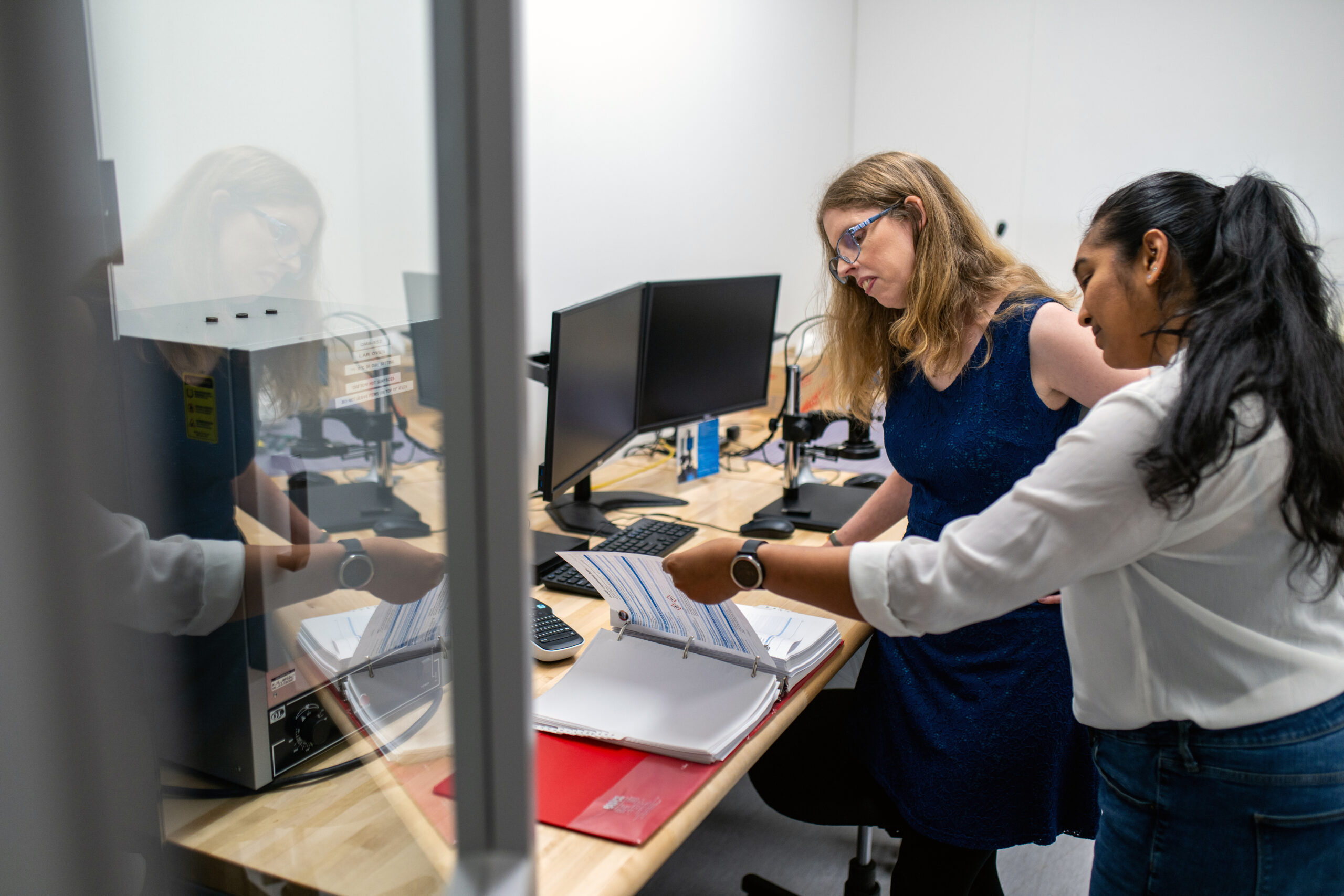 Two women working together in an office or lab setting. One woman, wearing a blue dress and safety glasses, sits at a desk with dual computer monitors, while the other woman, in a white blouse and jeans, stands beside her, pointing to documents in an open binder on the desk. The scene is viewed through a glass partition, with reflections partially visible, and the workspace includes technical equipment and office supplies.