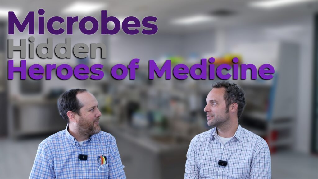 Two professionals in plaid shirts and microphones have a discussion in a laboratory setting. The text overlay reads "Microbes: Hidden Heroes of Medicine," emphasizing the role of microorganisms in medical advancements.