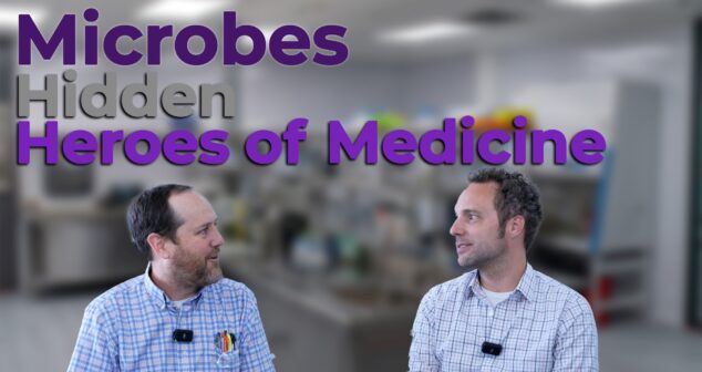 Two professionals in plaid shirts and microphones have a discussion in a laboratory setting. The text overlay reads "Microbes: Hidden Heroes of Medicine," emphasizing the role of microorganisms in medical advancements.