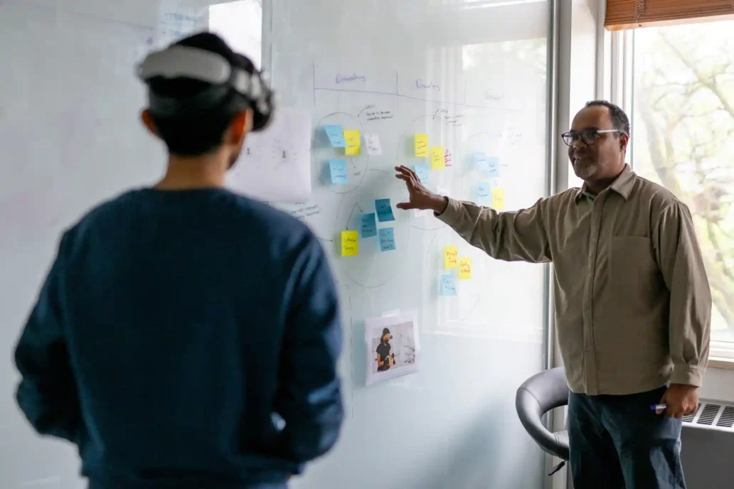 Two medtech professionals collaborating at a whiteboard covered in UX design notes and post-its, discussing user experience strategy for a medical device.