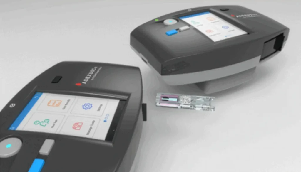 Two ADEXUSDx point-of-care analyzers with a test cassette shown between them on a clean white background.