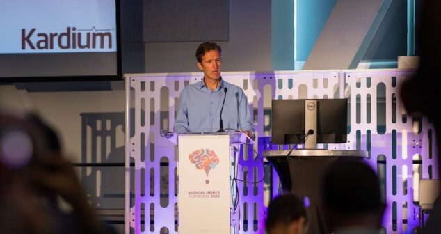 Kevin Chaplin wearing a light blue shirt stands at a podium, speaking into a microphone. The podium features a colorful brain-themed logo and the text "Medical Device Playbook 2024." Behind him is a screen displaying the logo "Kardium" and a modern backdrop with blue and purple lighting. An audience is partially visible in the foreground.