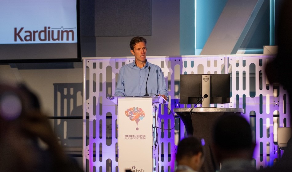 Kevin Chaplin wearing a light blue shirt stands at a podium, speaking into a microphone. The podium features a colorful brain-themed logo and the text "Medical Device Playbook 2024." Behind him is a screen displaying the logo "Kardium" and a modern backdrop with blue and purple lighting. An audience is partially visible in the foreground.