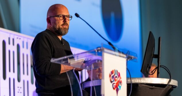 Andy Talbot, wearing a black sweater, speaks at a podium with a microphone. The podium features a colorful brain-themed logo associated with the "Medical Device Playbook 2024." A computer monitor is visible beside him, and the background displays a large blue presentation slide with abstract elements.