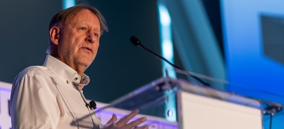 Glenn Bindley speaks at a podium with a microphone attached to his collar. The background features blue and white tones, likely from a stage setup.