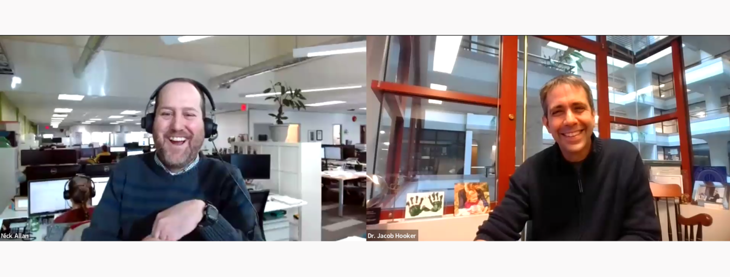 A screenshot from a virtual webinar or video call hosted by StarFish Medical. On the left, a man labeled 'Nick Allan' is wearing a headset and laughing, sitting in an open office space with multiple computer monitors and employees in the background. On the right, a man labeled 'Dr. Jacob Hooker' is smiling while sitting in an office with large windows, framed pictures, and artwork behind him.