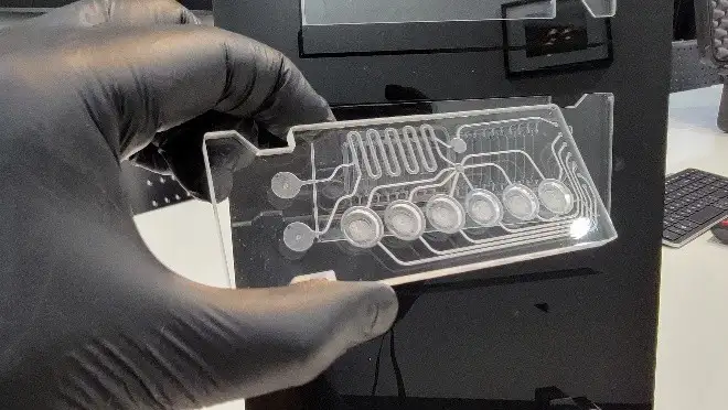 Gloved hand holding a transparent microfluidic cartridge with etched channel network and reservoir features