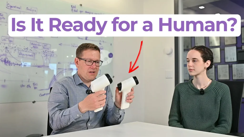 Two people seated at a table holding handheld medical device prototypes, with text overlay reading "Is It Ready for a Human?" — MedDevice by Design episode on clinical prototypes for human use