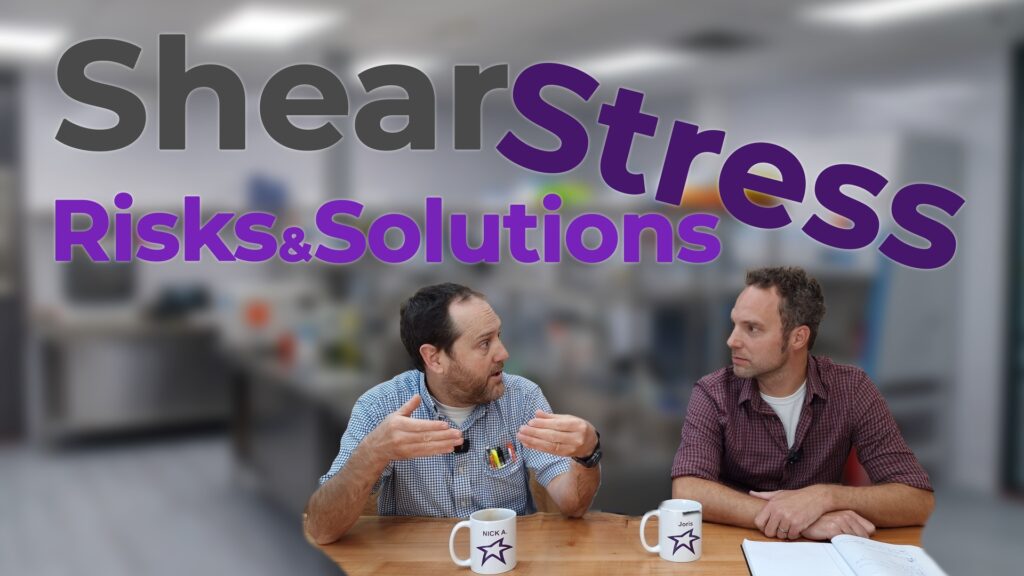 Two individuals sit at a wooden table in a bright laboratory setting, deeply engaged in discussion. One person gestures animatedly, emphasizing a point, while the other listens intently, with a notebook open in front of them. Both have mugs on the table labeled with their names, adding a personal touch. The text overlay reads 'Shear Stress: Risks & Solutions' in bold and engaging typography, with the backdrop of lab equipment creating a professional yet approachable atmosphere.