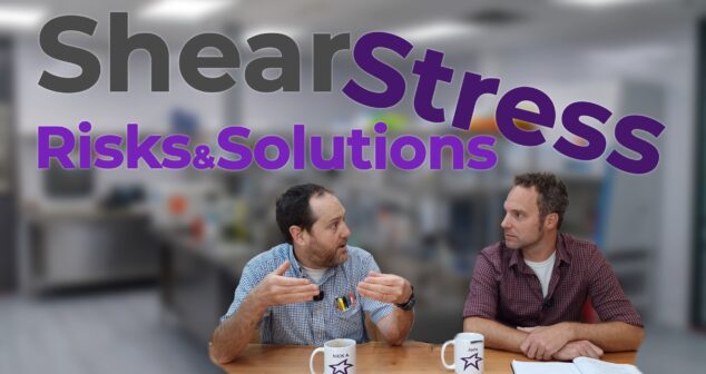 Two individuals sit at a wooden table in a bright laboratory setting, deeply engaged in discussion. One person gestures animatedly, emphasizing a point, while the other listens intently, with a notebook open in front of them. Both have mugs on the table labeled with their names, adding a personal touch. The text overlay reads 'Shear Stress: Risks & Solutions' in bold and engaging typography, with the backdrop of lab equipment creating a professional yet approachable atmosphere.