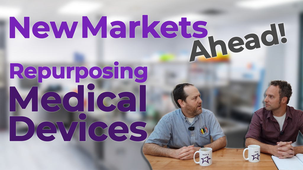 Two professionals discussing strategies for repurposing medical devices into new market sectors, sitting at a table with mugs featuring their names (Nick and Joris) and a star logo. The background is a blurred office or laboratory setting. The overlay text reads, 'New Markets Ahead! Repurposing Medical Devices,' in bold purple and black fonts, emphasizing innovation and market expansion.