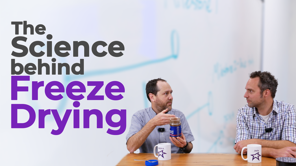 Two men sit at a table discussing the science behind freeze-drying. One of them holds a jar of freeze-dried coffee, illustrating the topic. The text overlay reads "The Science Behind Freeze Drying" in bold letters. A whiteboard with diagrams is visible in the background, reinforcing the technical discussion.