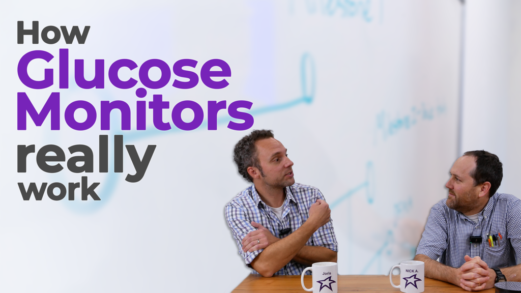 Two men sit at a table discussing glucose monitors, with the text "How Glucose Monitors Really Work" overlaid on the image. One man gestures while speaking, and both have coffee mugs labeled with their names. The background features a whiteboard with blurred writing.