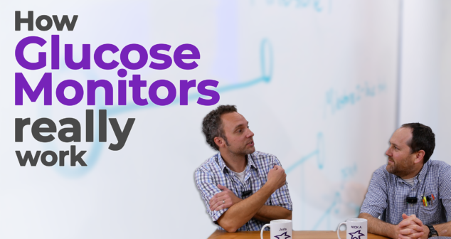 Two men sit at a table discussing glucose monitors, with the text "How Glucose Monitors Really Work" overlaid on the image. One man gestures while speaking, and both have coffee mugs labeled with their names. The background features a whiteboard with blurred writing.