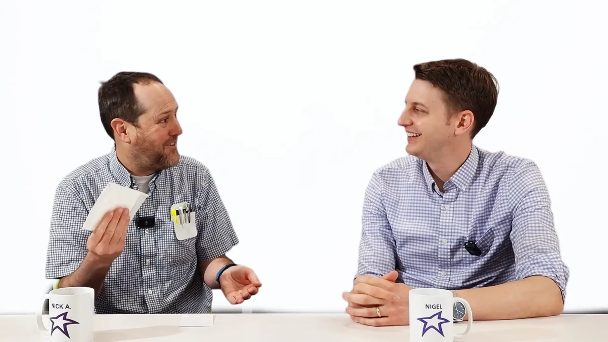 Two men, Nick A. (left) and Nigel (right), sit at a white table, engaging in a lively and friendly conversation. Both wear checkered shirts and lavalier microphones, suggesting a filmed discussion or interview. Nick holds tissue samples in one hand and gestures animatedly, while Nigel smiles in response. Each has a white mug labeled with their name and a purple star logo. The background is a bright white, creating a clean and professional studio setting.