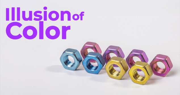 A row of metallic hex nuts anodized in vibrant colors including blue, pink, purple, and gold are arranged on a white surface. To the left, bold purple text reads "Illusion of Color," suggesting a theme about visual perception or surface treatment.
