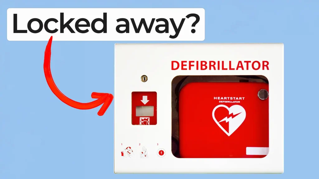A red defibrillator inside a white wall-mounted cabinet labeled DEFIBRILLATOR. A red arrow points to it, with overlay text reading ‘Locked away?’ in bold black letters on a white highlight.