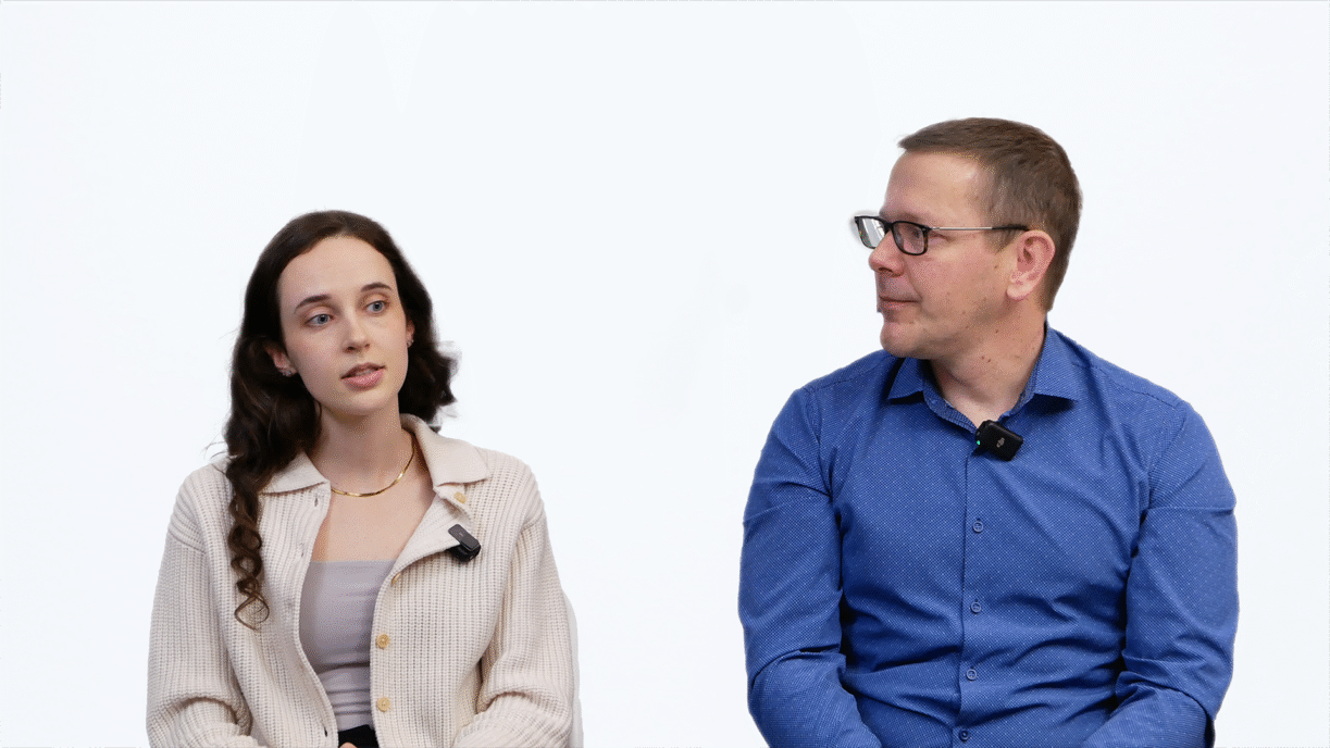 Ariana Wilson and Mark Drlik seated against a white background, wearing lavalier microphones. The woman on the left has long brown curly hair and is wearing a beige cardigan over a taupe top. The man on the right is wearing glasses and a blue button-up shirt. They appear to be engaged in conversation or a video interview.