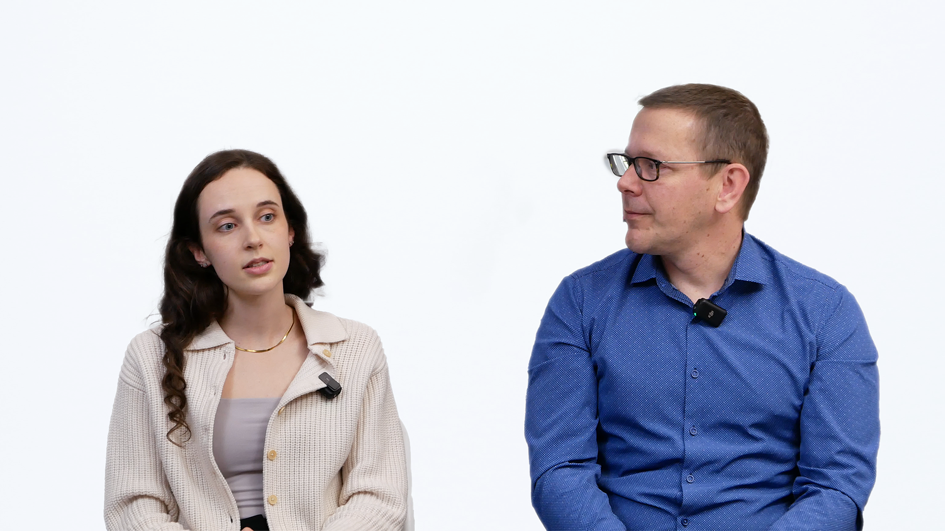 Ariana Wilson and Mark Drlik seated against a white background, wearing lavalier microphones. The woman on the left has long brown curly hair and is wearing a beige cardigan over a taupe top. The man on the right is wearing glasses and a blue button-up shirt. They appear to be engaged in conversation or a video interview.