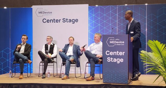 Panel discussion at the Silicon Valley MEDdevice event, with four seated panelists and one standing moderator on the Center Stage. The backdrop features geometric patterns and event branding, with a palm plant visible on the side of the stage.