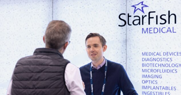 Two individuals engaged in a conversation at a StarFish Medical booth at MD&M West. The background displays the StarFish Medical logo along with text highlighting areas of expertise, including medical devices, diagnostics, biotechnology, microfluidics, imaging, optics, implantables, and ingestibles.