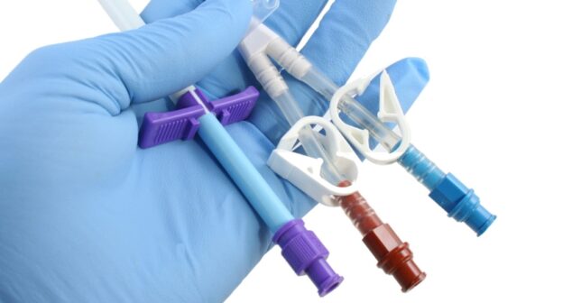 A gloved hand holding multiple medical catheters with various colored connectors and clamps. The catheters are flexible, hollow tubes designed for medical applications such as fluid drainage or medication delivery. The background is plain white, highlighting the details of the catheters.